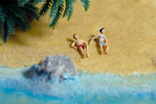 Miniature People Toy Figure Photography. Top Eagle View Of Men And Girl Couple Relaxing, Lying At Beach Sand When Daylight At Seaside