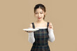 Happy beautiful Asian model holding a plate with copy space for separate products on beige background.