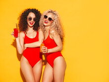Two Beautiful Sexy Smiling Hipster Women In Red Summer Swimwear Bathing Suits.Trendy Models With Afro Curls Hairstyle Having Fun In Studio.Hot Female Isolated On Yellow.Drinking Lemonade From Bottle