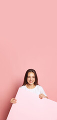 Vertival flyer with young pretty girl in white t-shirt holding huge empty sheet of paper isolated on pink background. Concept of beauty, art, fashion, youth and emotions