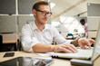 © Mediaphotos - Content creative man with tattoo sitting at table in office of marketing company and editing presentation on laptop