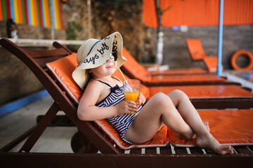 Cute child girl in wide-brimmed hat and sunglasses in swimsuit on wooden sun lounger orange, summer vacation with children