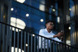 © Mediaphotos - Serious young Afro-American businessman in glasses standing by railing against glassy building facade and answering sms on phone