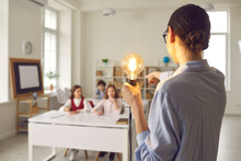 Innovative Learning, Creative Educational Study For Graduation And School Student Success Concept. Rear View On Teacher Holding Glowing Idea Lightbulb Pointing At Pupil Sitting At Classroom Desk