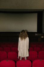 Curly-haired Girl Stands Facing The Cinema Screen Between Rows Of Red Chairs