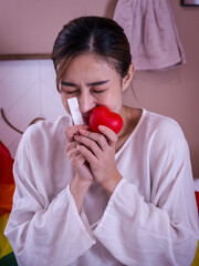 Young lesbian enjoys pregnancy test, holding a red heart and the tube with rainbow flag on background.