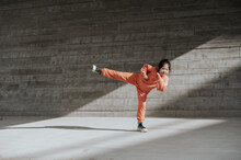 Young Girl In Workout Clothes Does A High Kick By A Concrete Wall.