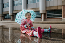 Young Girl Sits In A Puddle With Rain Boots And An Umbrella.