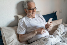 Senior Man With Vitiligo Lying In Bed Looking At Mobile Phone
