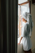 © Berena Alvarez/Stocksy - Young woman in white bathrobe take air at the window
