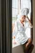 © Berena Alvarez/Stocksy - Young woman in white bathrobe take air at the window