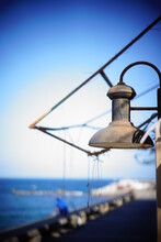 Close-up Of Old And Worn Fisherman's Wharf Lamp, With Blue Sky In The Background And Sea With Out Of Focus People Fishing In The Background, Nostalgia Concept, Fishermen,
