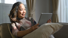 Retired Woman Sitting On Comfortable Sofa And Checking Email, News Online On Tablet. Elderly Technology Concept