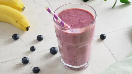 Wall Mural - Blueberry and banana smoothie in a glass with a straw on white table. Summer refreshing fruit drink. 4k video