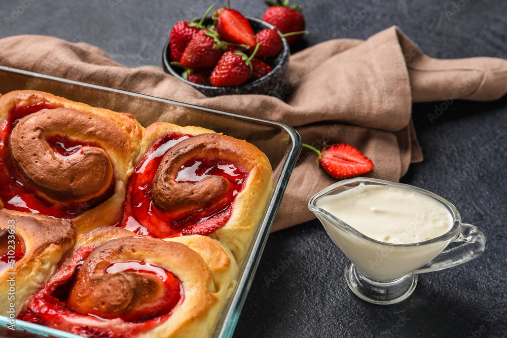 Baking dish with strawberry cinnamon rolls, cream and napkin on dark background, closeup