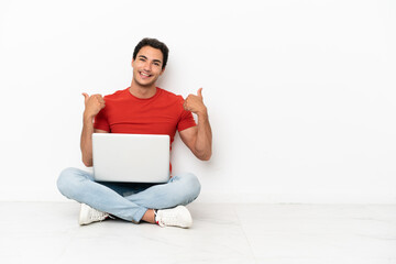 Wall Mural - Caucasian handsome man with a laptop sitting on the floor with thumbs up gesture and smiling