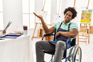 Wall Mural - Young african american artist man sitting on wheelchair at art studio smiling cheerful presenting and pointing with palm of hand looking at the camera.