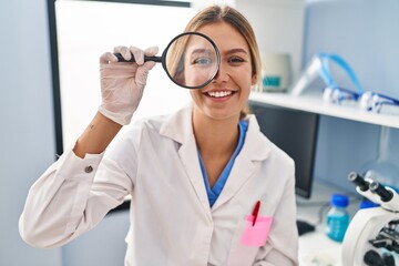 Sticker - Young blonde woman working at scientist laboratory using magnifying glass looking positive and happy standing and smiling with a confident smile showing teeth