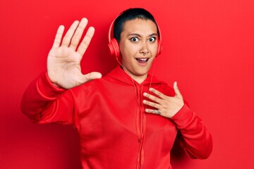 Beautiful hispanic woman with short hair listening to music using headphones afraid and terrified with fear expression stop gesture with hands, shouting in shock. panic concept.