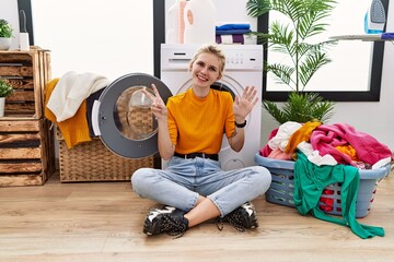 Sticker - Young blonde woman doing laundry sitting by washing machine showing and pointing up with fingers number seven while smiling confident and happy.