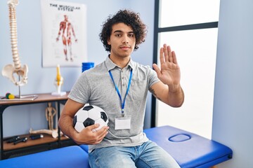 Canvas Print - Hispanic man with curly hair working as football physiotherapist doing stop sing with palm of the hand. warning expression with negative and serious gesture on the face.