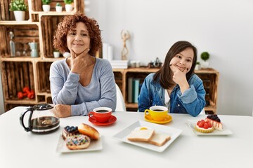 Canvas Print - Family of mother and down syndrome daughter sitting at home eating breakfast looking confident at the camera with smile with crossed arms and hand raised on chin. thinking positive.