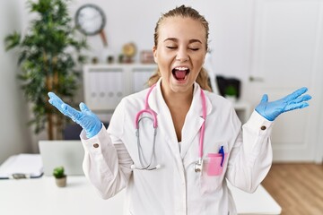 Poster - Young caucasian woman wearing doctor uniform and stethoscope at the clinic celebrating mad and crazy for success with arms raised and closed eyes screaming excited. winner concept