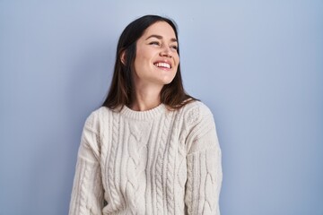 Poster - Young brunette woman standing over blue background looking away to side with smile on face, natural expression. laughing confident.