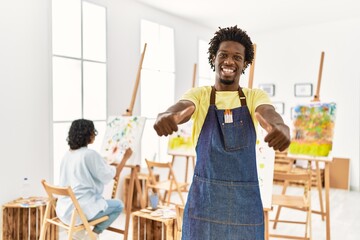 Wall Mural - African young man standing at art studio approving doing positive gesture with hand, thumbs up smiling and happy for success. winner gesture.