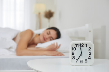 Close up of white plastic alarm clock set for 7 AM on bedside table in bedroom, with happy peaceful tranquil young woman sleeping on bed in blurred background. Sleep, morning, daily routine concepts