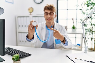 Canvas Print - Young caucasian doctor man wearing doctor uniform and using stethoscope at the clinic smiling happy pointing with hand and finger