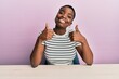 Young african american woman wearing casual clothes sitting on the table success sign doing positive gesture with hand, thumbs up smiling and happy. cheerful expression and winner gesture.
