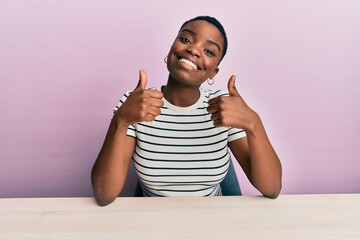 Sticker - Young african american woman wearing casual clothes sitting on the table success sign doing positive gesture with hand, thumbs up smiling and happy. cheerful expression and winner gesture.