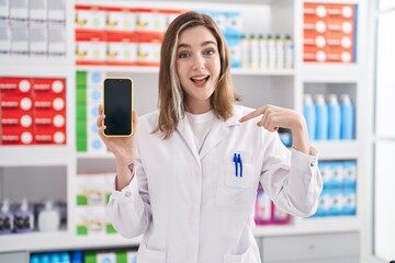 Poster - Blonde caucasian woman working at pharmacy drugstore showing smartphone screen pointing finger to one self smiling happy and proud