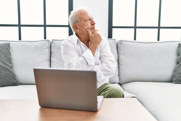 Wall Mural - Senior man using laptop at home sitting on the sofa looking confident at the camera with smile with crossed arms and hand raised on chin. thinking positive.