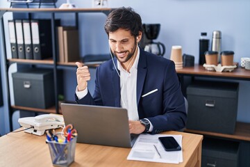 Canvas Print - Handsome latin man working at the office using laptop pointing thumb up to the side smiling happy with open mouth