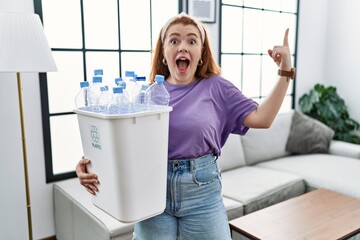 Poster - Young redhead woman holding recycling wastebasket with plastic bottles smiling amazed and surprised and pointing up with fingers and raised arms.