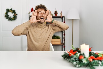 Wall Mural - Young handsome man with beard sitting on the table by christmas decoration smiling cheerful playing peek a boo with hands showing face. surprised and exited