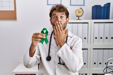 Sticker - Young hispanic doctor man holding support green ribbon at clinic covering mouth with hand, shocked and afraid for mistake. surprised expression