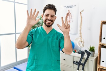 Poster - Young man with beard working at pain recovery clinic showing and pointing up with fingers number eight while smiling confident and happy.