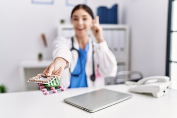 Young hispanic doctor woman wearing doctor uniform holding prescription pills at clinic surprised with an idea or question pointing finger with happy face, number one