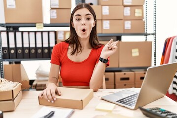 Poster - Young hispanic woman preparing order working at storehouse surprised pointing with finger to the side, open mouth amazed expression.