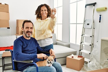 Poster - Middle age hispanic couple smiling happy. Man sitting on wheelchair with dog on his legs and woman holding key of new home.