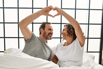 Canvas Print - Middle age hispanic couple smiling happy making heart symbol with hands. Sitting on the bed at home.