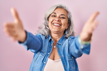 Sticker - Middle age woman with grey hair standing over pink background looking at the camera smiling with open arms for hug. cheerful expression embracing happiness.