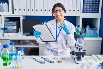 Sticker - Hispanic young woman working at scientist laboratory cheerful with a smile on face pointing with hand and finger up to the side with happy and natural expression