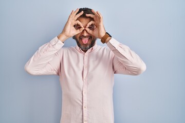 Sticker - Handsome middle age man wearing elegant shirt background doing ok gesture like binoculars sticking tongue out, eyes looking through fingers. crazy expression.