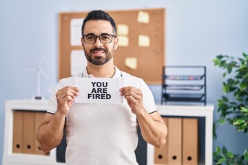 Sticker - Young hispanic man with beard holding you are fired banner at the office smiling with a happy and cool smile on face. showing teeth.