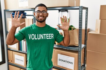 Poster - Young indian man volunteer holding donations box showing and pointing up with fingers number ten while smiling confident and happy.