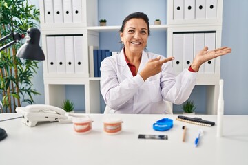 Sticker - Hispanic mature dentist woman working at the clinic amazed and smiling to the camera while presenting with hand and pointing with finger.